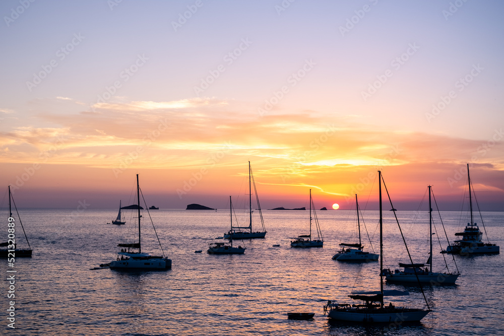Several yachts and boats anchored near the coast relax watching the sunset on the horizon of the Mediterranean islands.