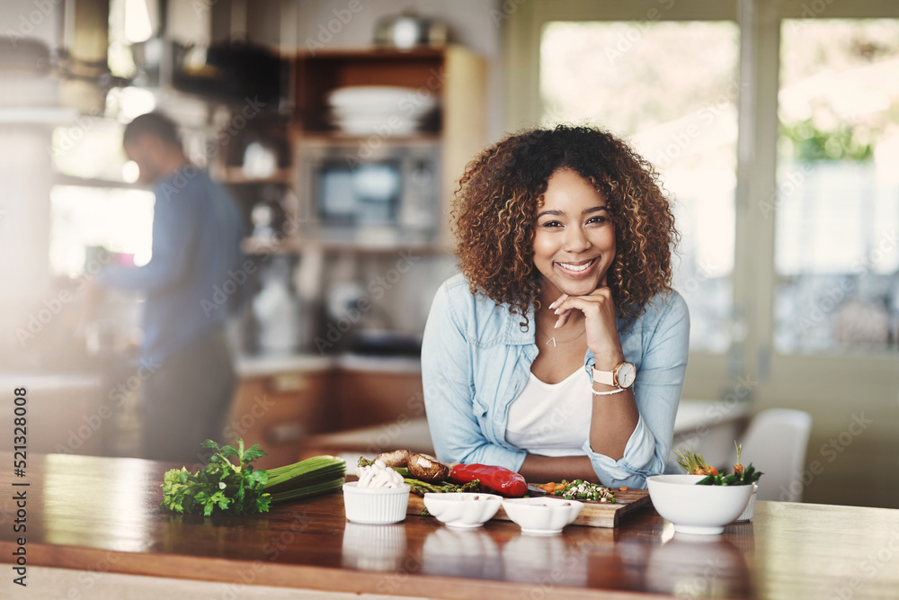 Portrait of a happy, healthy and carefree young woman preparing a healthy meal at home with her husband in the background. Black wife making an organic vegetarian salad for lunch in a kitchen