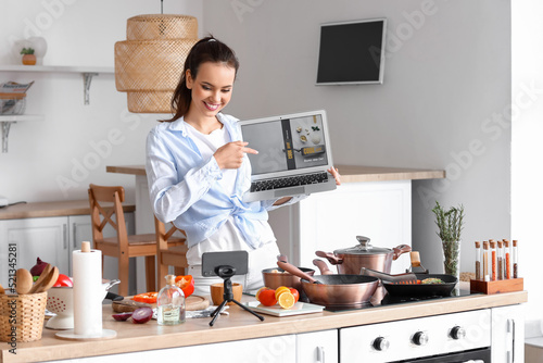 Young woman with laptop recording cooking video tutorial in kitchen