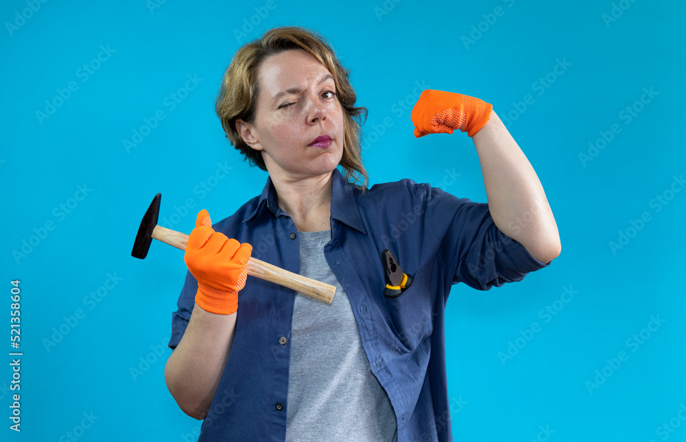 A strong woman in a shirt and orange gloves holds a hammer in her hand on a blue isolated background. Portrait of a female builder in a working shirt A strong woman in a shirt and orange gloves holds a hammer in her hand on a blue isolated background. Portrait of a female builder in a working shirt