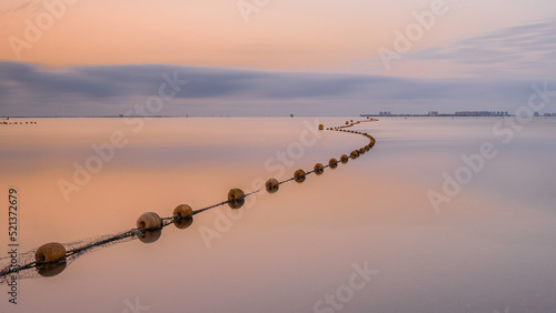 A string of buoys, stretching to the horizon of a very calm, mirror like sea, on a tranquil summer morning, in the Mediterranean. The sky is orange as the sun comes up. 