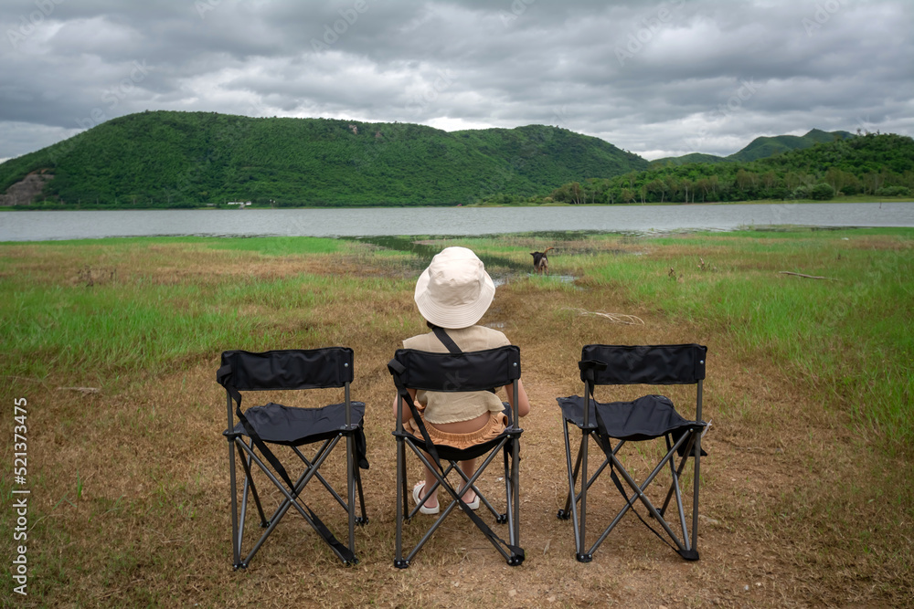 The young woman sits on the camping chair and looks at the view of the reservoir at the Huai Phak Reservoir, Tha Yang District, Phetchaburi, Thailand. The young woman sits on the camping chair and looks at the view of the reservoir at the Huai Phak Reservoir, Tha Yang District, Phetchaburi, Thailand.