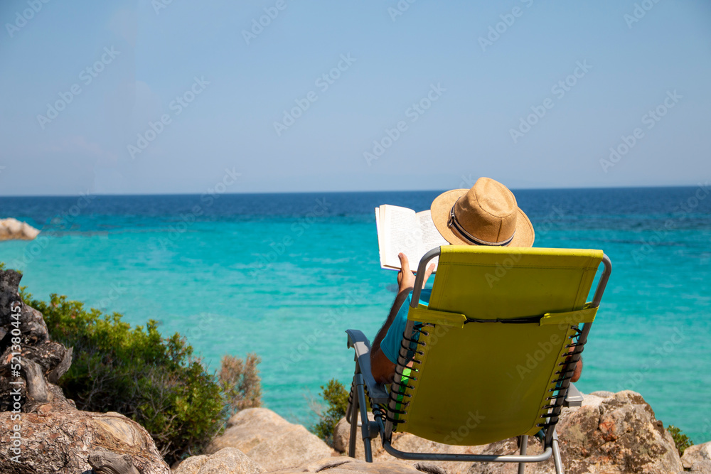 Fototapeta premium Portrait of a handsome man lying on a deckchair and reading a book. Happy mature man next to the beach on Aegean sea with beautiful view. Summer vacation concept.