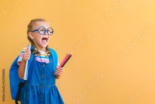 emotional excited surprise face schoolgirl child in glasses in school uniform holds colored pencils, notebook, backpack open mouth to empty copy space attentive isolated on yellow background