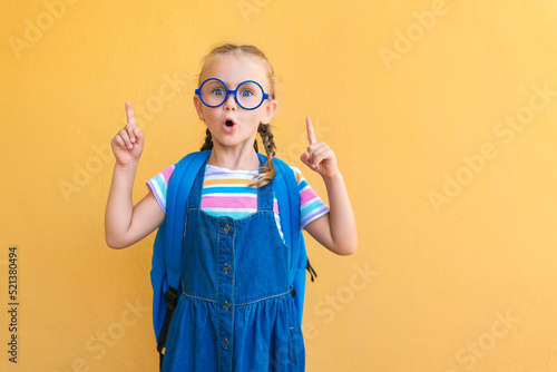 emotional excited surprise face schoolgirl child in glasses in school blue uniform with backpack school bag open mouth point fingers up to empty copy space attentive isolated on yellow background