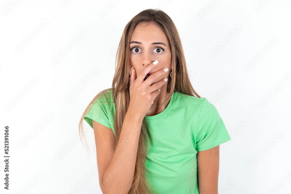 Emotional young beautiful woman wearing green T-shirt over white background gasps from astonishment, covers opened mouth with palm, looks shocked at camera. Emotional young beautiful woman wearing green T-shirt over white background gasps from astonishment, covers opened mouth with palm, looks shocked at camera.