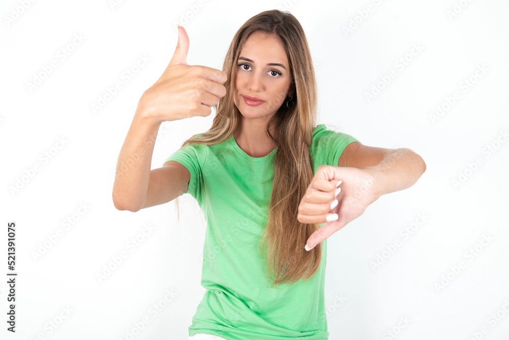 young beautiful woman wearing green T-shirt over white background showing thumbs up and thumbs down, difficult choose concept young beautiful woman wearing green T-shirt over white background showing thumbs up and thumbs down, difficult choose concept