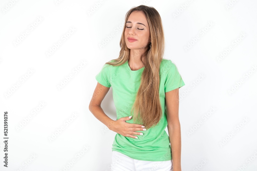 young beautiful woman wearing green T-shirt over white background touches tummy, smiles gently, eating and satisfaction concept. young beautiful woman wearing green T-shirt over white background touches tummy, smiles gently, eating and satisfaction concept.