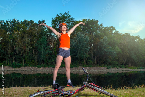 girl cyclist in a helmet in the morning sits on the banks of the river and rejoices in nature