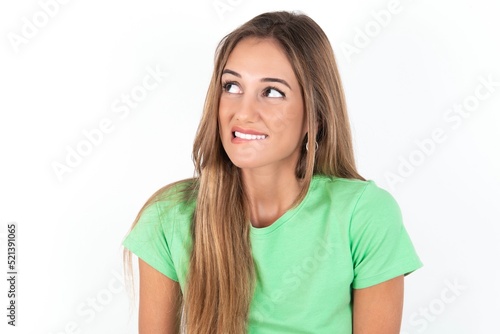 Amazed young beautiful woman wearing green T-shirt over white background bitting lip and looking tricky to empty space.