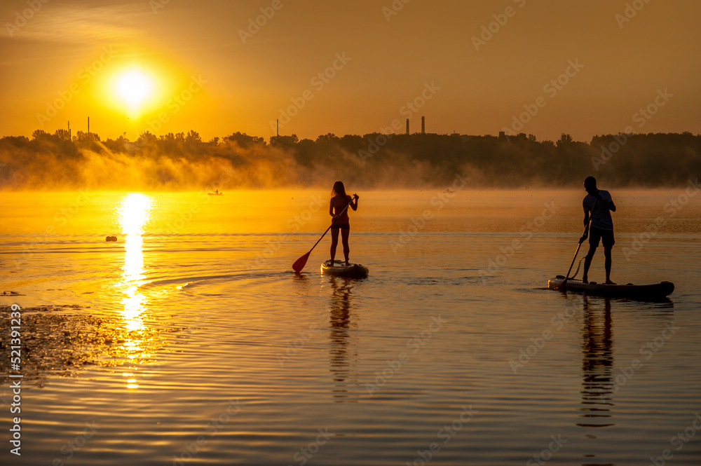 silhouettes of a couple on SUP boards on the water of the lake, red sky with morning fog, active recreation