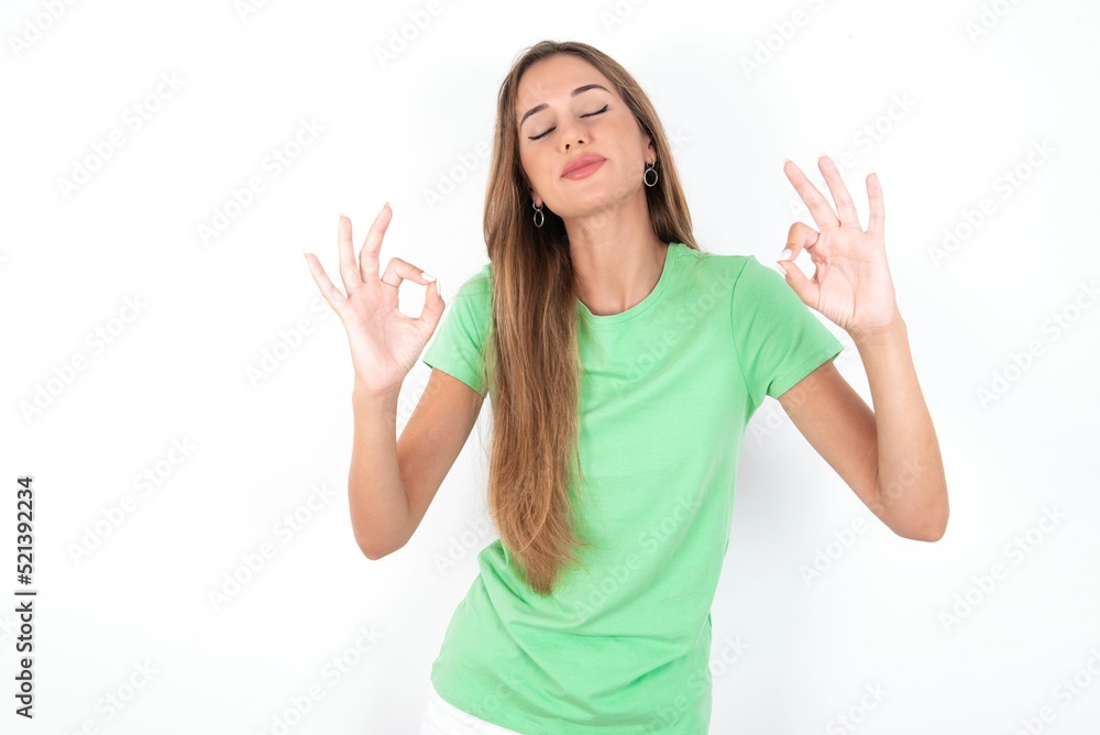 young beautiful woman wearing green T-shirt over white background, relax and smiling with eyes closed doing meditation gesture with fingers. Yoga concept. young beautiful woman wearing green T-shirt over white background, relax and smiling with eyes closed doing meditation gesture with fingers. Yoga concept.