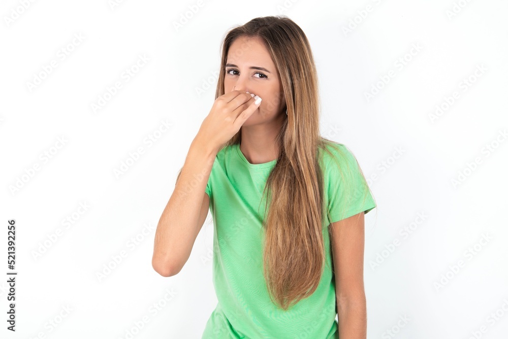 young beautiful woman wearing green T-shirt over white background, smelling something stinky and disgusting, intolerable smell, holding breath with fingers on nose. Bad smell young beautiful woman wearing green T-shirt over white background, smelling something stinky and disgusting, intolerable smell, holding breath with fingers on nose. Bad smell