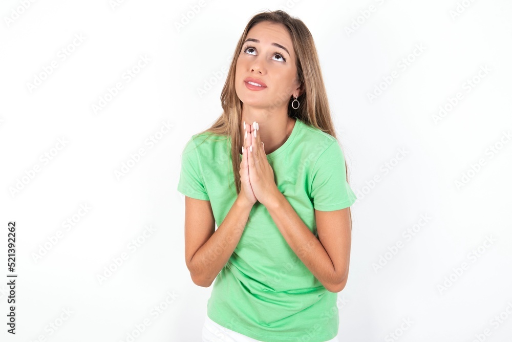 young beautiful woman wearing green T-shirt over white background, begging and praying with hands together with hope expression on face very emotional and worried. Please God young beautiful woman wearing green T-shirt over white background, begging and praying with hands together with hope expression on face very emotional and worried. Please God