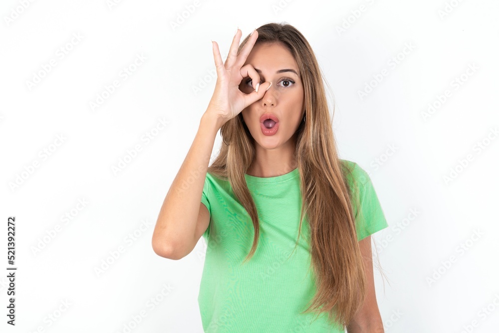 young beautiful woman wearing green T-shirt over white background, doing ok gesture shocked with surprised face, eye looking through fingers. Unbelieving expression. young beautiful woman wearing green T-shirt over white background, doing ok gesture shocked with surprised face, eye looking through fingers. Unbelieving expression.