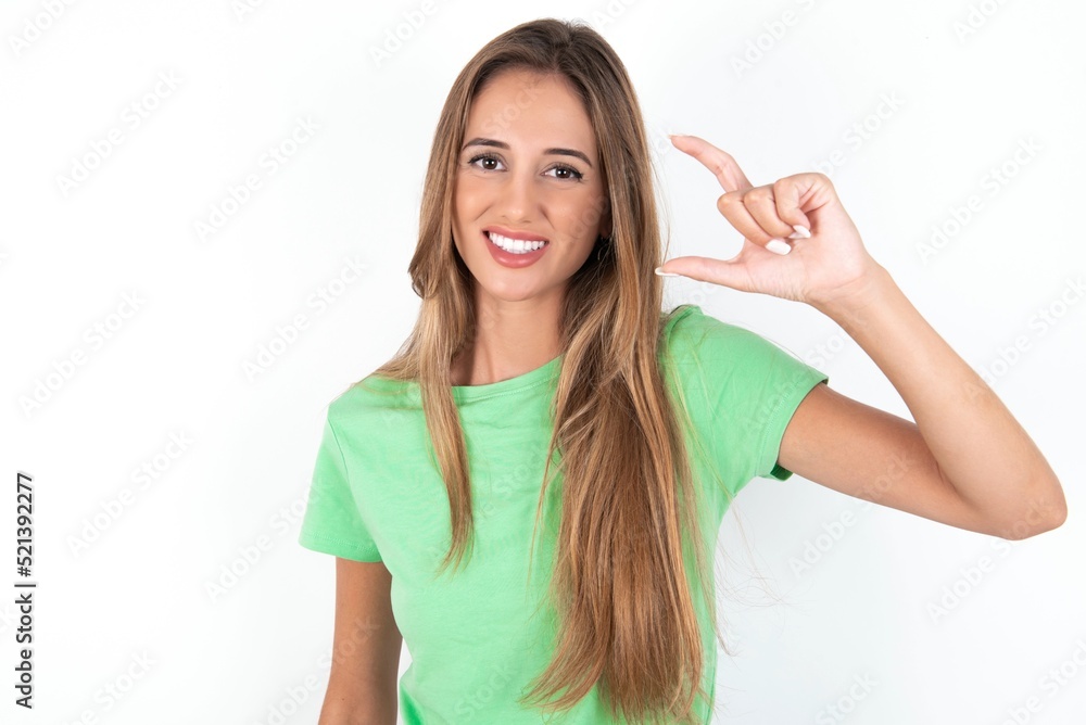 young beautiful woman wearing green T-shirt over white background, smiling and confident gesturing with hand doing small size sign with fingers looking and the camera. Measure concept young beautiful woman wearing green T-shirt over white background, smiling and confident gesturing with hand doing small size sign with fingers looking and the camera. Measure concept