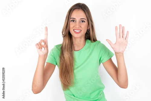 young beautiful woman wearing green T-shirt over white background, showing and pointing up with fingers number six while smiling confident and happy.