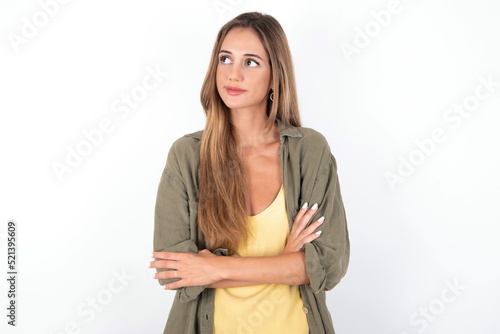Charming thoughtful young beautiful woman wearing green overshirt over white background stands with arms folded concentrated somewhere with pensive expression thinks what to do