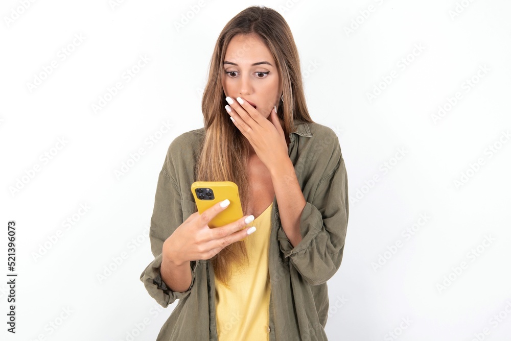young beautiful woman wearing green overshirt over white background being deeply surprised, stares at smartphone display, reads shocking news on website, Omg, its horrible! young beautiful woman wearing green overshirt over white background being deeply surprised, stares at smartphone display, reads shocking news on website, Omg, its horrible!