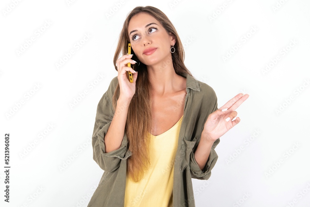 young beautiful woman wearing green overshirt over white background speaks on mobile phone spends free time indoors calls to friend. young beautiful woman wearing green overshirt over white background speaks on mobile phone spends free time indoors calls to friend.