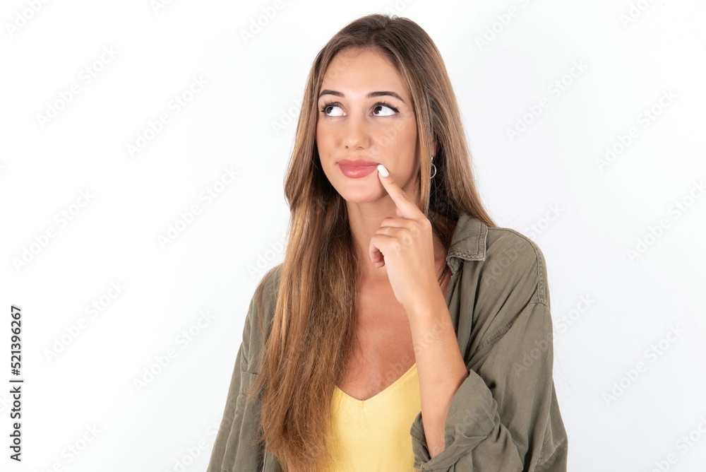 Lovely dreamy young beautiful woman wearing green overshirt over white background keeps finger near lips looks aside copy space. Lovely dreamy young beautiful woman wearing green overshirt over white background keeps finger near lips looks aside copy space.
