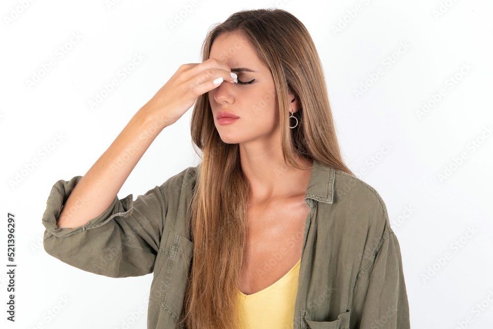 Sad young beautiful woman wearing green overshirt over white background suffering from headache holding hand on her face Sad young beautiful woman wearing green overshirt over white background suffering from headache holding hand on her face