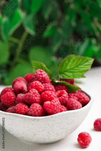 Bowl of fresh ripe raspberries with green leaves on white table against blurred background, closeup. Space for text