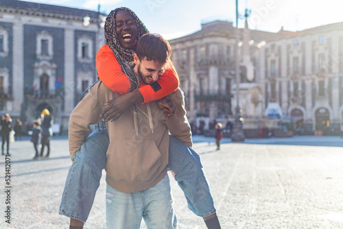 Smiling diverse young boyfriend carrying girlfriend on his back and laughing outdoors - boyfriend giving piggyback ride to girlfriends - Young multiracial couple having piggyback – happy multi ethnic 
