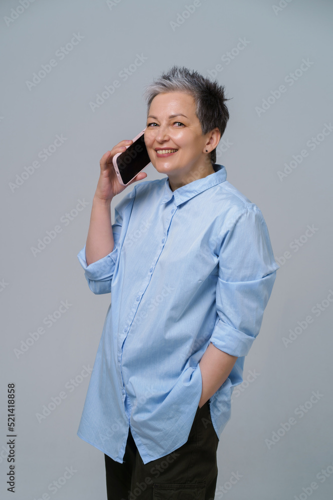 Mature woman with grey hair talking holding smartphone at her ear looking sideways, smiling wearing long blue shirt. Pretty woman in blue blouse isolated on white background Mature woman with grey hair talking holding smartphone at her ear looking sideways, smiling wearing long blue shirt. Pretty woman in blue blouse isolated on white background