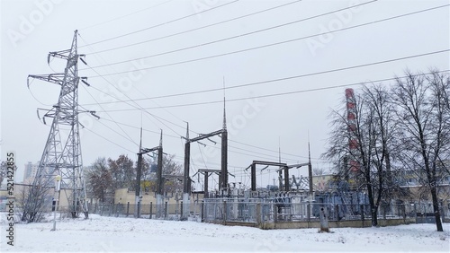 A transformer substation with a metal power line tower, ceramic insulators, and lightning rods is located on a snow-covered lawn and surrounded by a fence. Nearby are a factory chimney and buildings. 