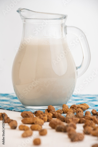 Close-up of glass pitcher with horchata and tiger nuts on white table with blue cloth, selective focus, white background, vertical