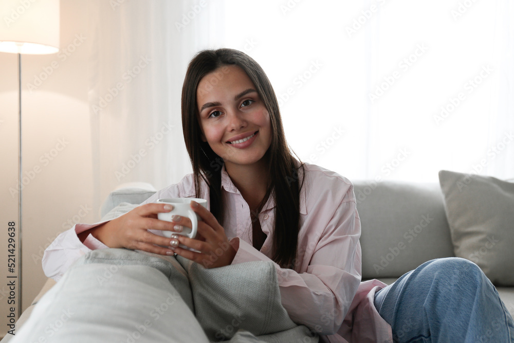 Portrait of young beautiful woman wearing a beige knitted sweater sitting on the couch holding a big cup of coffee. Brown eyed female with smiling facial expression. Copy space, background, close up Portrait of young beautiful woman wearing a beige knitted sweater sitting on the couch holding a big cup of coffee. Brown eyed female with smiling facial expression. Copy space, background, close up