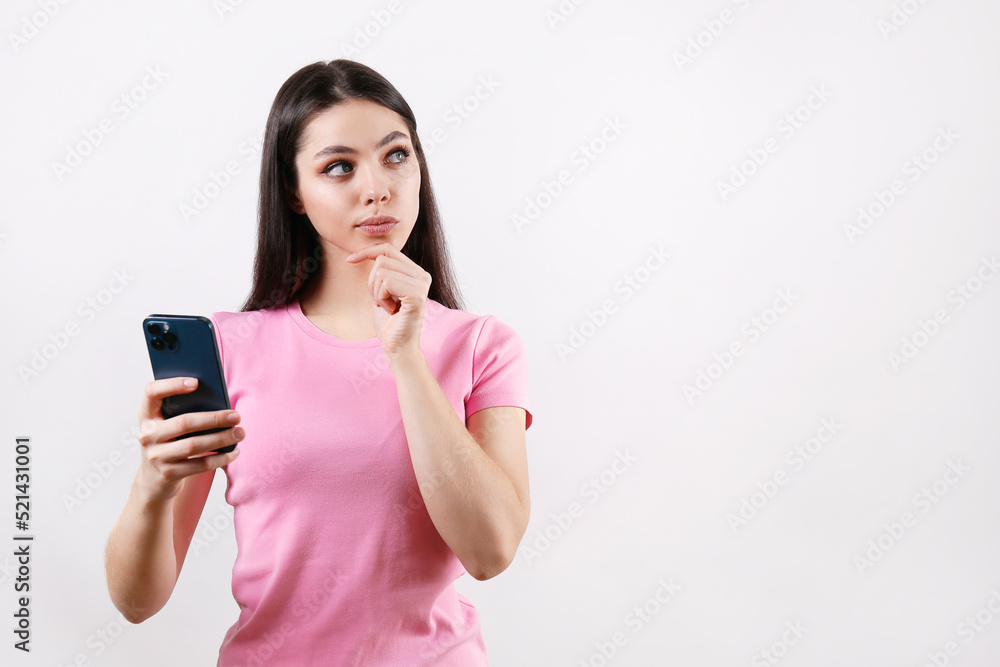 Young beautiful woman with thoughtful facial expression holding a blank screen phone. Portrait of female model with cellphone calculating in her mind. Close up, copy space, isolated white background. Young beautiful woman with thoughtful facial expression holding a blank screen phone. Portrait of female model with cellphone calculating in her mind. Close up, copy space, isolated white background.
