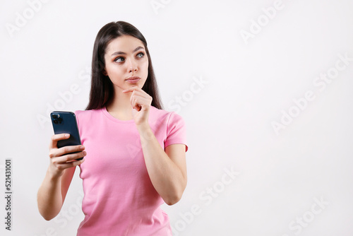 Young beautiful woman with thoughtful facial expression holding a blank screen phone. Portrait of female model with cellphone calculating in her mind. Close up, copy space, isolated white background.