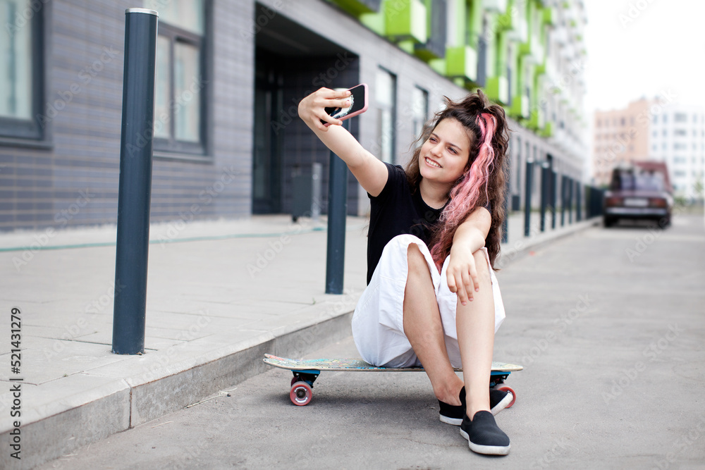 Fototapeta premium Teen girl sitting on road on skateboard, taking selfie on her phone and smiling. Lifestyle youth culture. Trendy teen with long pink colored hair