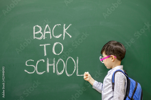 cute little boy preschooler is writing on blackboard with white chalk the words back to school.kid is wearing funny round party glasses with no glass inside.education,learning,classroom interior