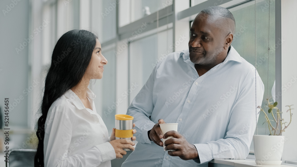 Two diverse age multi-ethnic colleagues businesspeople co-workers partners friends african senior adult man arab young woman standing indoors talk drink coffee tea have pleasant informal conversation Two diverse age multi-ethnic colleagues businesspeople co-workers partners friends african senior adult man arab young woman standing indoors talk drink coffee tea have pleasant informal conversation