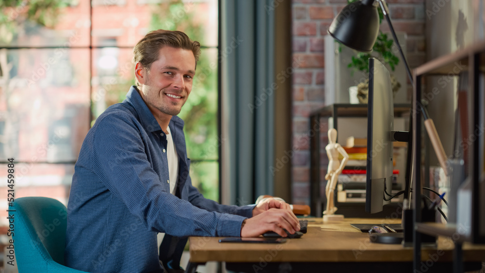 Portrait of a Young Handsome Man Sitting at Home, Working on Desktop Computer. Creative Male Looking Directly at Camera and Smiles. Living Room with Big Window in Loft Apartment. Portrait of a Young Handsome Man Sitting at Home, Working on Desktop Computer. Creative Male Looking Directly at Camera and Smiles. Living Room with Big Window in Loft Apartment.