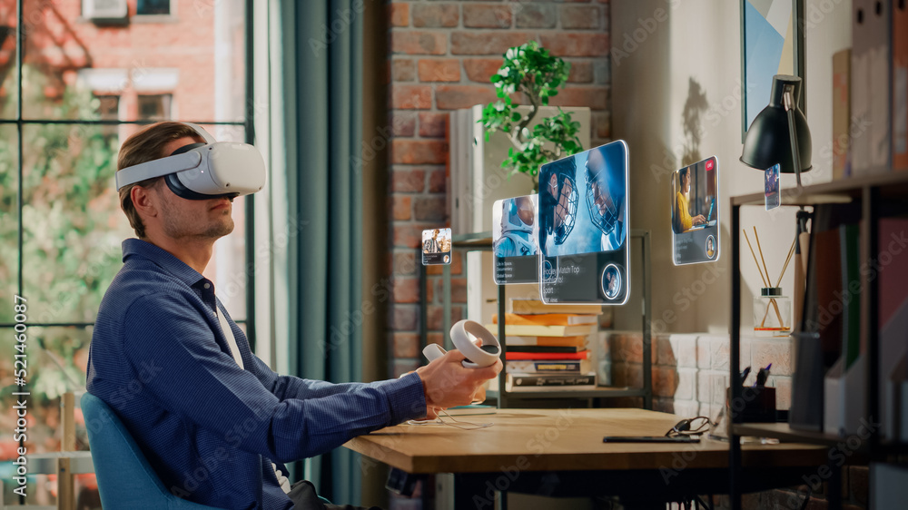 Excited Young Man Sitting in Loft Living Room at Home, Using Virtual Reality Headset with Controllers to Check Social Media Streaming Application. Male Choosing to Watch American Football Highlights. Excited Young Man Sitting in Loft Living Room at Home, Using Virtual Reality Headset with Controllers to Check Social Media Streaming Application. Male Choosing to Watch American Football Highlights.