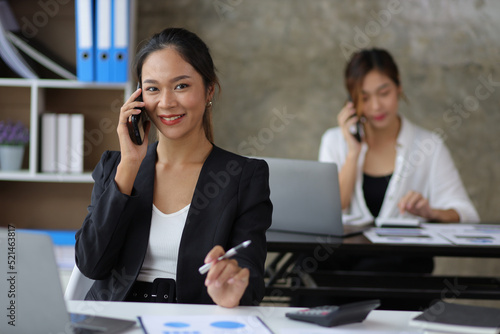 Attractive Asian businesswoman talking on mobile phone working in modern office. Happy business woman talking on mobile phone while analyzing monthly work schedule in laptop.