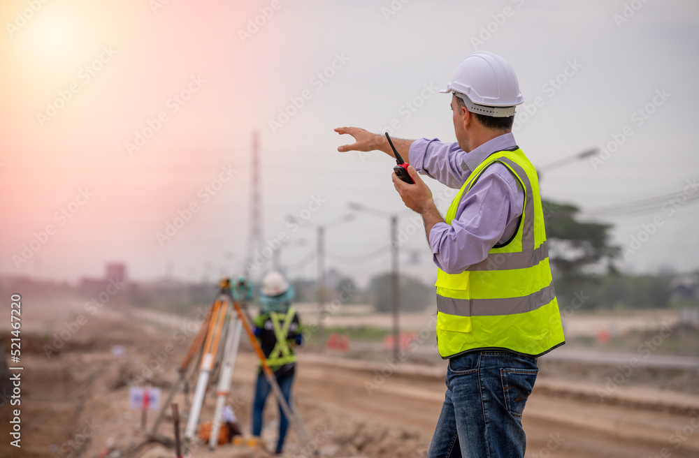 Surveyor engineer wearing safety uniform ,helmet and radio communication with equipment theodolite to measurement positioning on the construction site of the road with construct machinery background Surveyor engineer wearing safety uniform ,helmet and radio communication with equipment theodolite to measurement positioning on the construction site of the road with construct machinery background