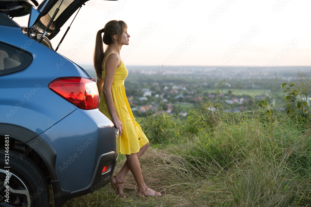 Yong happy female driver resting near her car enjoying sunset view of summer nature. Travel and recreation concept