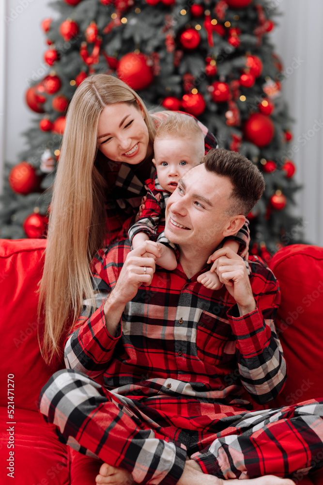 The theme of the family holiday is New year and Christmas. Young European family in the same pyjamas: mom, dad, baby boy are sitting on the red sofa by the festive Christmas tree on Christmas evening