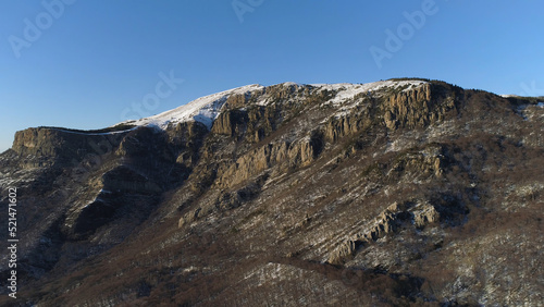 Aerial view of mountains on a sunny autumn day on blue clear sky background. Shot. Flying over snowy steep rocky walls with growing bald trees.