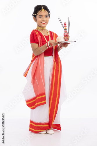 Portrait of beautiful bengali girl holding puja thali with conch shell, sindoor dani and incense sticks in traditional sari
 photo