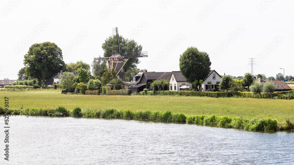 Fototapeta premium Windmill and houses along the river De Linge near the Dutch village of Zoelen in the Betuwe.