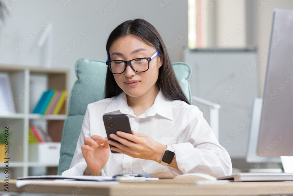 A young beautiful Asian business woman in glasses is holding a phone in her hands, dialing a message. Sitting at a desk in a modern office, looking at the camera A young beautiful Asian business woman in glasses is holding a phone in her hands, dialing a message. Sitting at a desk in a modern office, looking at the camera