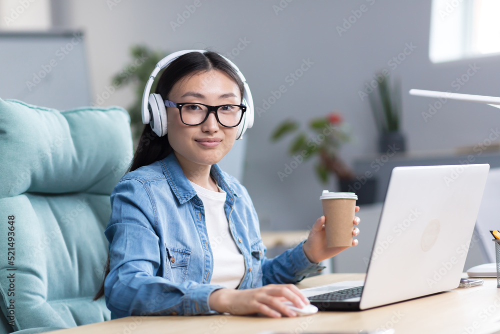 Online training. Young beautiful Asian student girl in glasses studies at a laptop in headphones, drinks coffee. Learns remotely, sits at a desk in a modern office, looks at the camera. Online training. Young beautiful Asian student girl in glasses studies at a laptop in headphones, drinks coffee. Learns remotely, sits at a desk in a modern office, looks at the camera.
