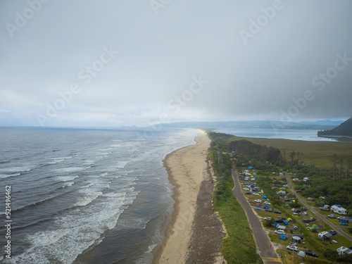 Shooting from the air. Small town on the green hilly coast of the ocean. White foamy waves crash against the sandy shore. Ecology, environmental protection, tourism.