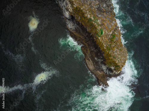 View from above. A small stone island covered with green moss in the middle of the ocean. Turquoise ocean water and white foamy waves wash the island from all sides. Abstraction.